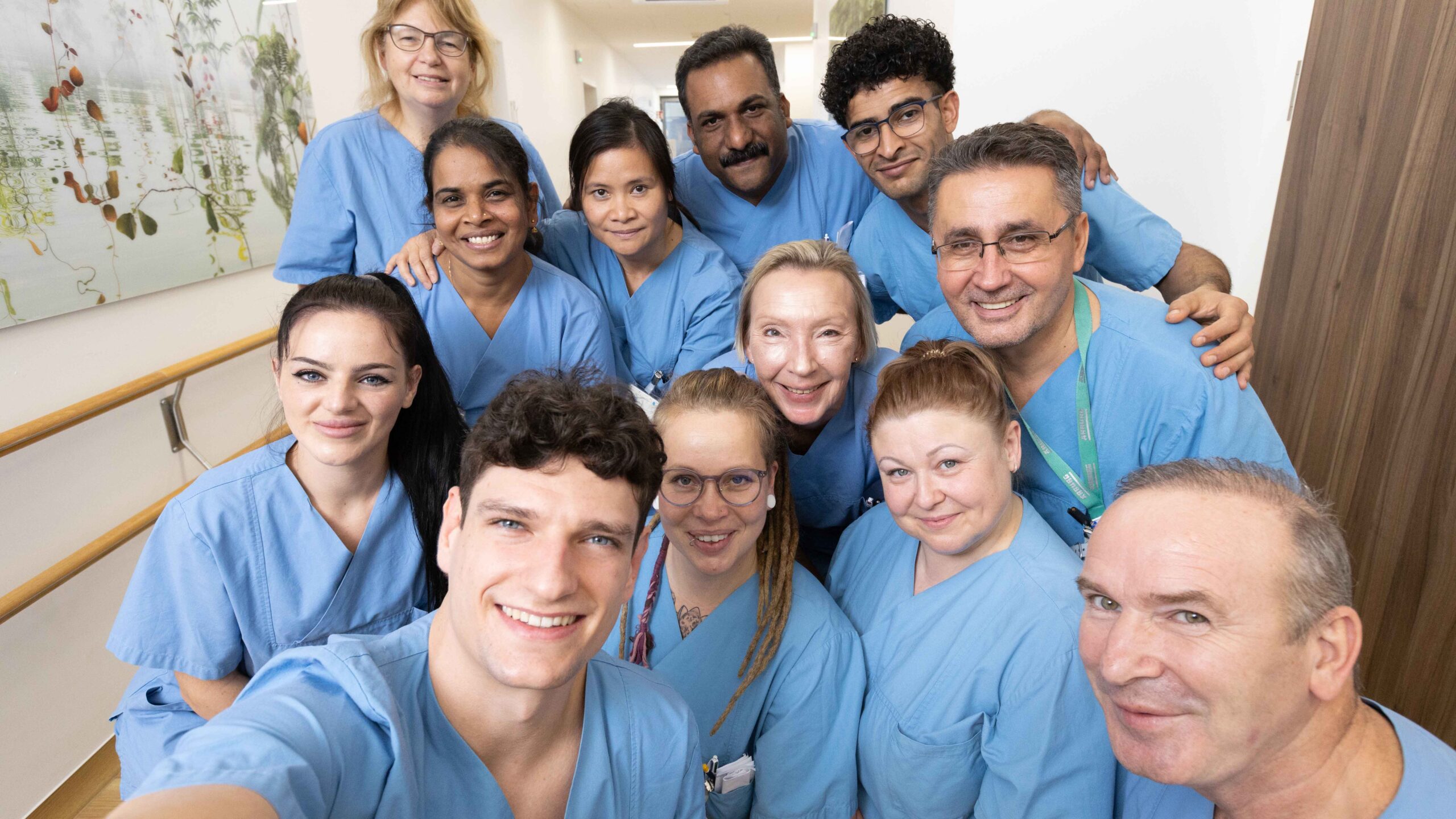 Ein Team vom zwölf Gesundheits- und Krankenpfleger*innen von einer Pfleges in blauer Arbeitskleidung. Sie stehen als Gruppe zusammmen und machen ein Selfie mit einem Smartphone